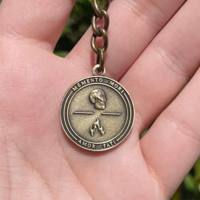 May include: A bronze-toned circular pendant with the inscriptions "MEMENTO MORI" and "AMOR FATI". The pendant displays a skull, a horizontal line, and a flame symbol. It is connected to a bronze chain.