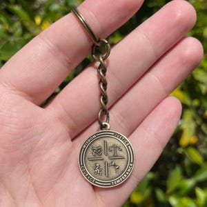May include: A bronze-coloured keyring with a round pendant featuring symbols and the words "Wisdom," "Justice," "Temperance," and "Courage." The pendant has a detailed design with a key ring and a twisted chain.