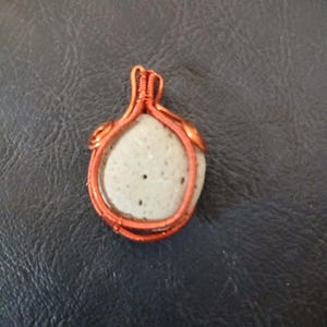 May include: A handcrafted pendant featuring a light gray stone wrapped in copper wire. The wire is intricately woven around the stone, creating a unique and organic design. The stone has small dark spots and a smooth texture. The pendant is displayed on a dark background.