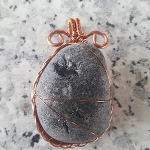 May include: A gray stone pendant wrapped in copper wire with a braided design. The pendant has a unique, natural texture.