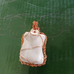 Puede incluir: Un colgante hecho a mano con una piedra blanca lisa envuelta en alambre de cobre. El alambre está intrincadamente tejido alrededor de la piedra, creando un borde decorativo y un lazo para una cadena. El colgante se muestra sobre un fondo verde.