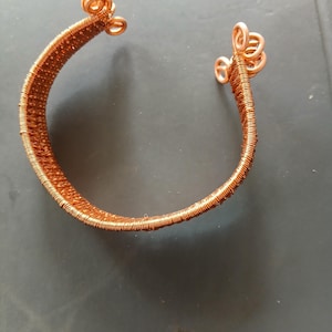 Könnte beinhalten: Kupferfarbenes Drahtarmband. Das Armband besteht aus eng gewickeltem Draht mit dekorativen Schlaufen an den Enden. Das Design ist schlicht und elegant, für den Alltag geeignet. Das Armband hat eine warme, metallische Farbe.