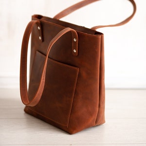 May include: A brown leather tote bag with a front pocket and two handles. The bag has a rectangular shape and is made of a smooth, textured leather. The handles are attached with silver rivets.