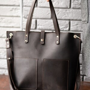May include: A brown leather tote bag with two front pockets and a long shoulder strap. The bag has gold hardware and stitching.