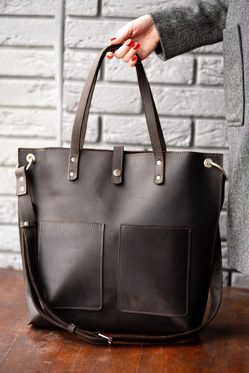 May include: A brown leather tote bag with two front pockets and a long shoulder strap. The bag has gold hardware and stitching.