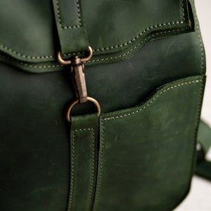 May include: A dark green leather backpack with a flap closure and a metal buckle. The backpack has a front pocket and a strap with a metal buckle.