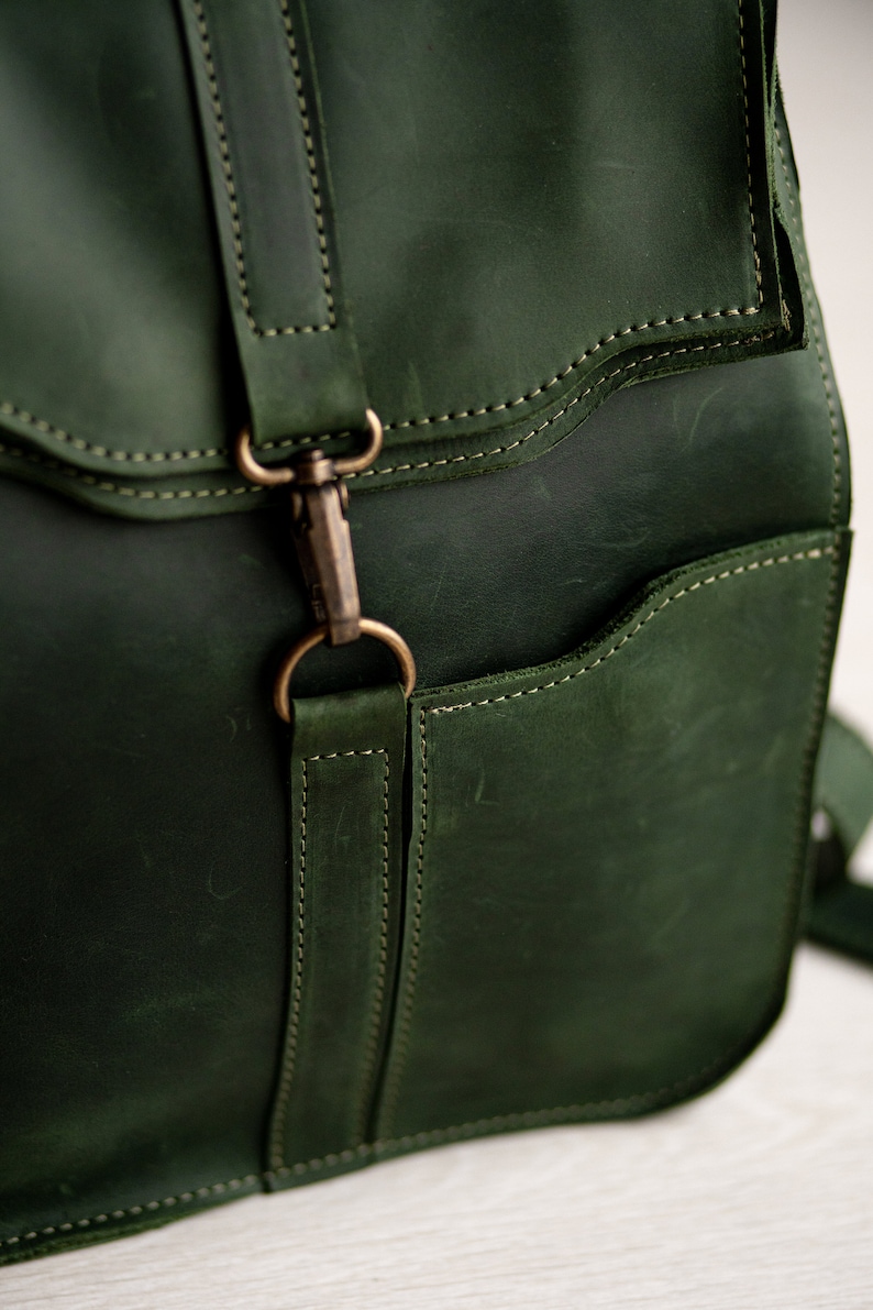 May include: A dark green leather backpack with a flap closure and a metal buckle. The backpack has a front pocket and a strap with a metal buckle.