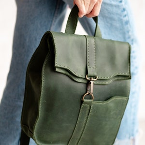 May include: A dark green leather backpack with a flap closure and a front pocket. The backpack has a metal buckle and a strap.