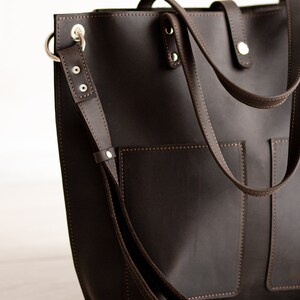 May include: A brown leather tote bag with a long strap and two pockets on the front. The bag has silver hardware and stitching.