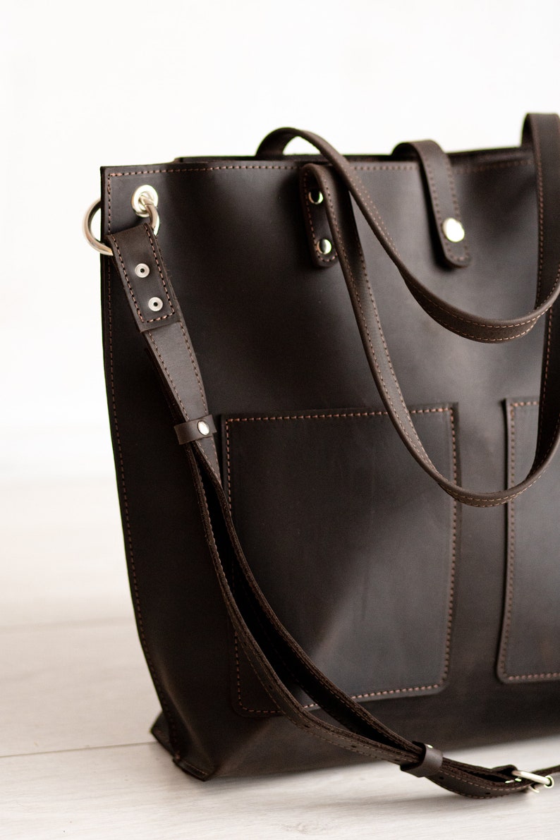 May include: A brown leather tote bag with a long strap and two pockets on the front. The bag has silver hardware and stitching.