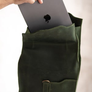 May include: A dark green leather backpack with a zippered pocket and a flap closure. A laptop computer is being placed inside the backpack.