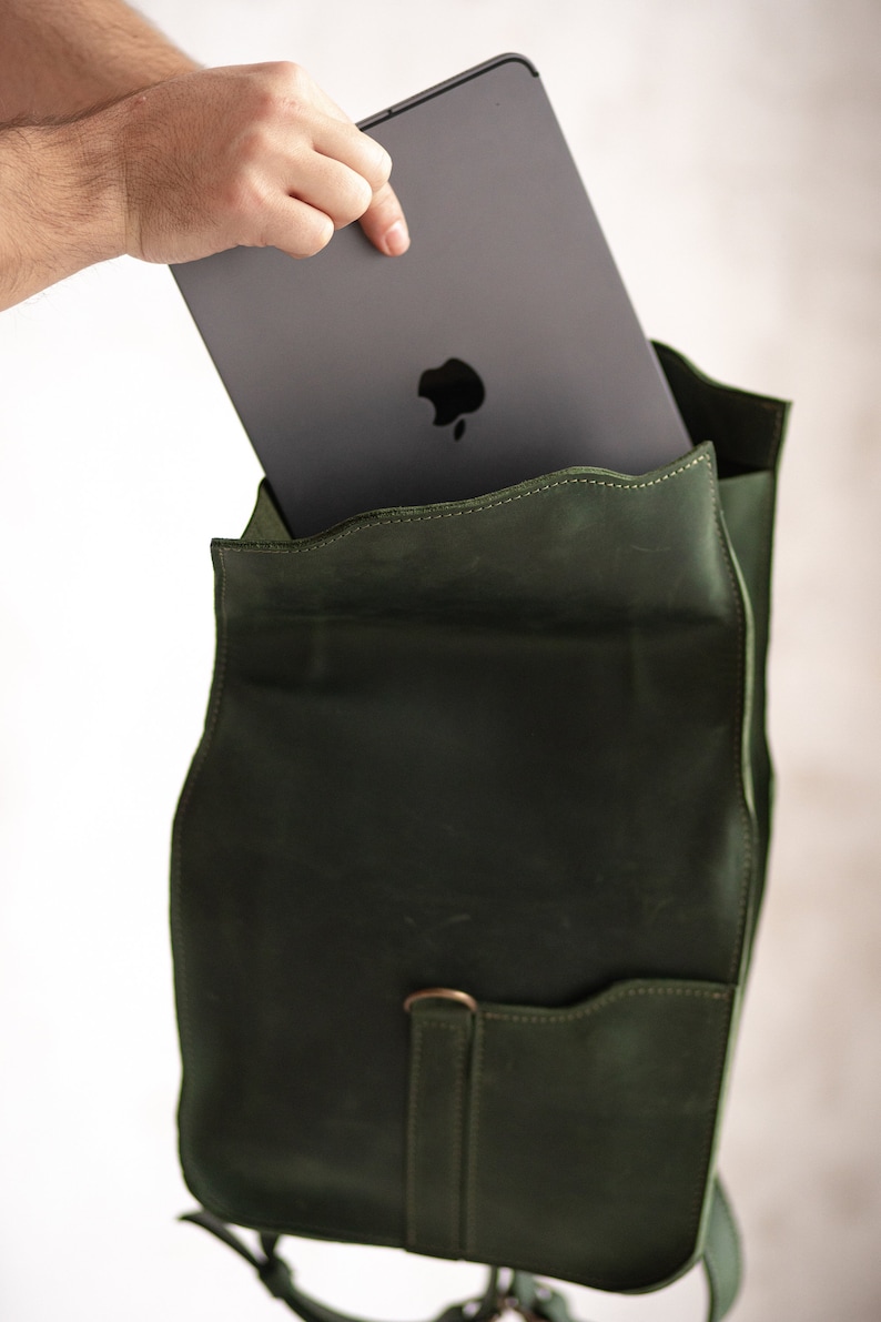 May include: A dark green leather backpack with a zippered pocket and a flap closure. A laptop computer is being placed inside the backpack.