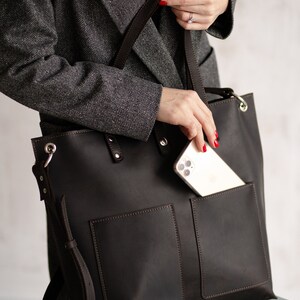 May include: A brown leather tote bag with a large pocket on the front and a smaller pocket on the side. The bag has a long strap that can be worn over the shoulder. A smartphone is visible in the front pocket.