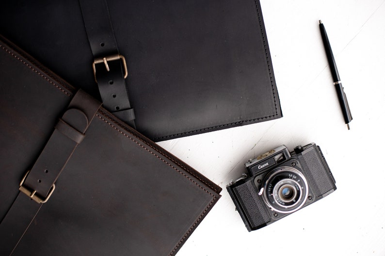May include: Two brown leather portfolios with straps and buckles. The portfolios are on a white surface with a black pen and a vintage camera.