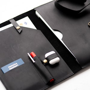 Puede incluir: Portafolio de cuero negro con cierre de solapa, correa y hebilla. El portafolio tiene varios bolsillos y compartimentos para llevar documentos, bolígrafos y otros artículos. El texto "UKRAINE PASSPORT" es visible en un pasaporte azul dentro del portafolio.