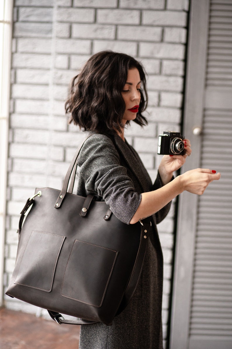 May include: A woman wearing a gray blazer holds a vintage camera in one hand and a large brown leather tote bag with two front pockets in the other hand.