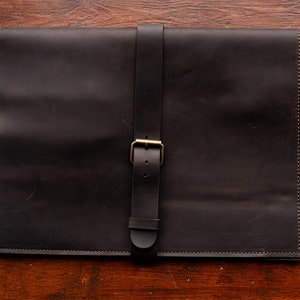 May include: A dark brown leather portfolio with a strap and a brass buckle closure.