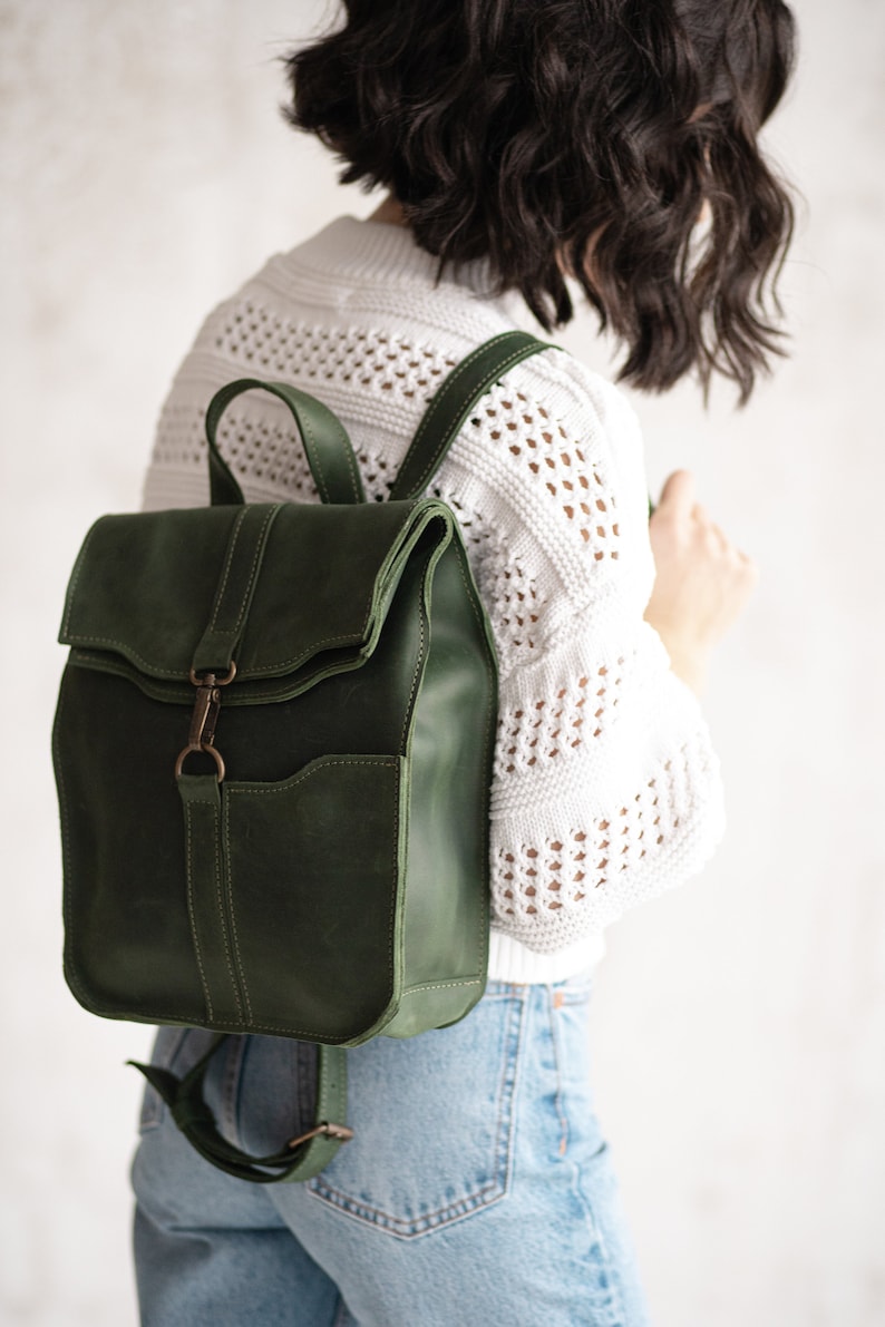 May include: A dark green leather backpack with a flap closure and a front pocket. The backpack has adjustable straps and a metal buckle.