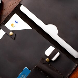 May include: A brown leather portfolio with a laptop sleeve, a pocket for a phone or passport, and a strap to secure the contents. The portfolio is open and the laptop sleeve is visible.