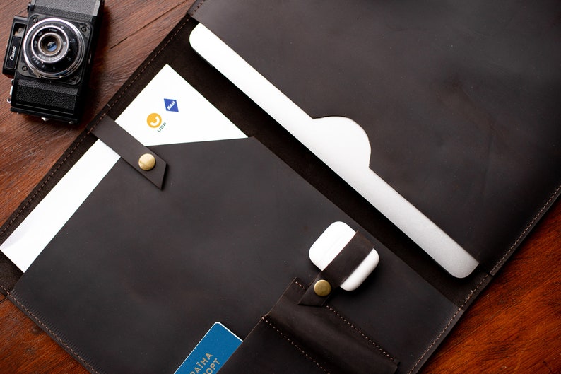 May include: A brown leather portfolio with a laptop sleeve, a pocket for a phone or passport, and a strap to secure the contents. The portfolio is open and the laptop sleeve is visible.