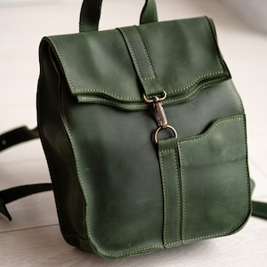 May include: A dark green leather backpack with a flap closure and a metal buckle. The backpack has a small pocket on the front and adjustable straps.
