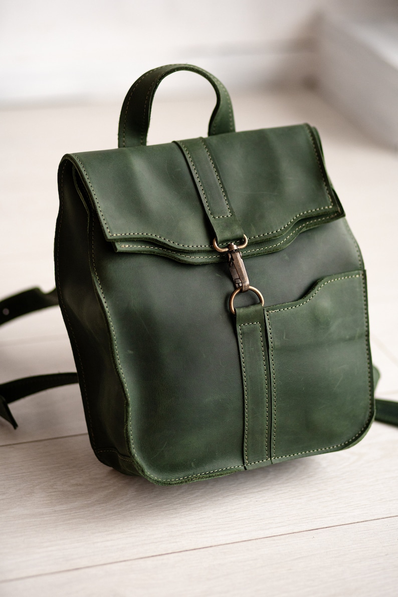 May include: A dark green leather backpack with a flap closure and a metal buckle. The backpack has a small pocket on the front and adjustable straps.