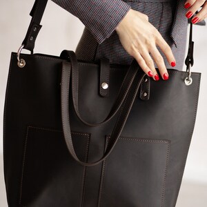 May include: A brown leather tote bag with two front pockets and a long strap. The bag is worn over the shoulder of a person wearing a patterned blouse and jeans.