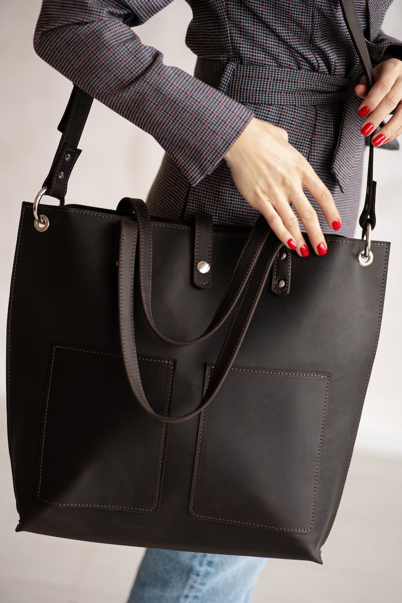 May include: A brown leather tote bag with two front pockets and a long strap. The bag is worn over the shoulder of a person wearing a patterned blouse and jeans.