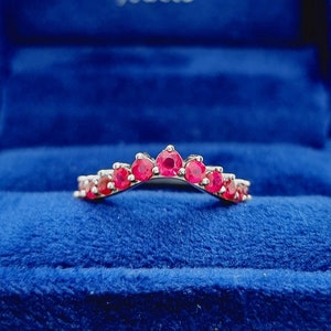 Könnte beinhalten: Ein zarter, gebogener Ehering mit einer Reihe von rosafarbenen Rubin-Steinen, die in eine silberne Band gefasst sind. Der Ring wird in einer blauen Samt-Ringbox präsentiert.