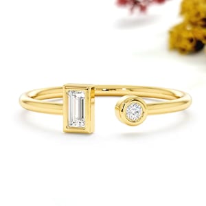 May include: A gold open ring with a rectangular and a round, clear gemstone. The ring is set against a white background with dried flowers in the upper right corner. The ring's design is modern and minimalist.