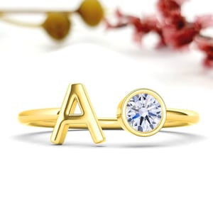 May include: A gold ring with the letter "A" and a clear, round gemstone. The ring is set against a white backdrop with blurred floral elements. The ring's design is simple and elegant, suitable for everyday wear.