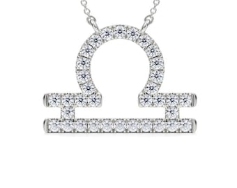 May include: A silver-colored Libra zodiac pendant necklace. The pendant is encrusted with small, clear gemstones, forming the Libra symbol. The necklace chain is also silver-colored and delicate. The pendant is centered on a white background.