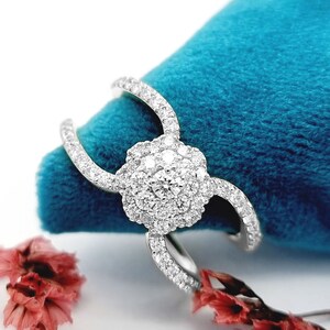 May include: A silver ring with a floral design, featuring a cluster of small, clear stones at the centre and along the curved bands. The ring is set against a teal background with dried pink flowers.