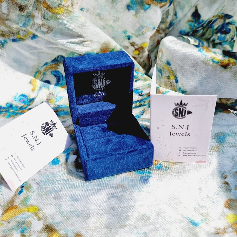 May include: A blue velvet ring box with a white label that says "S.N.J Jewels" and a crown logo. The box is open and empty.