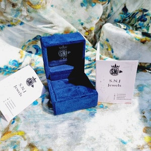 May include: A blue velvet ring box with a white label that says "S.N.J Jewels" and a crown logo. The box is open and empty.