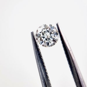 May include: A clear round diamond gemstone held by silver tweezers against a white background.