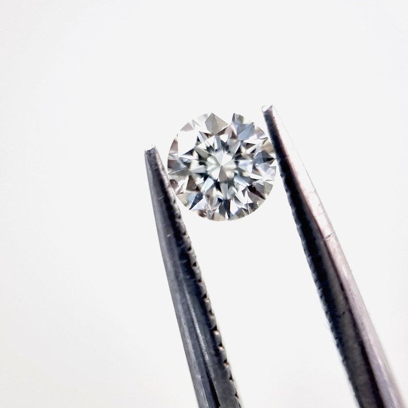 May include: A clear round diamond gemstone held by silver tweezers against a white background.