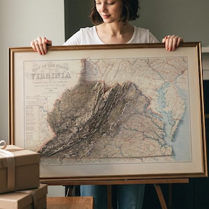 Op de afbeelding: Een ingelijste kaart van Virginia, met de titel "Map of the State of Virginia". De kaart heeft een 3D-reliëfeffect, dat de topografie van de staat laat zien. De lijst is goudkleurig. Twee cadeau dozen met linten staan op de voorgrond.