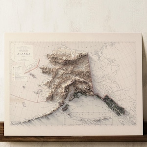 May include: A detailed relief map of Alaska, showing the state's topography in shades of brown and beige. The map includes geographical details and is presented on cream-coloured paper, mounted on a dark wooden shelf.