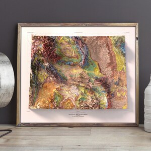 Puede incluir: Un mapa topogr&aacute;fico enmarcado de Wyoming con un dise&ntilde;o 3D texturizado y colorido. El mapa est&aacute; impreso en papel blanco y tiene un borde rosa claro. El marco es de madera y tiene un acabado marr&oacute;n claro.