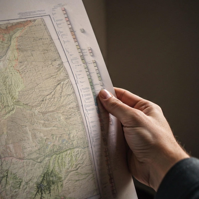 May include: A detailed vintage map with a hand holding the edge, revealing a portion of the map's intricate design. The map features a variety of colors, including greens, browns, and tans, and appears to be a topographical representation of a region.