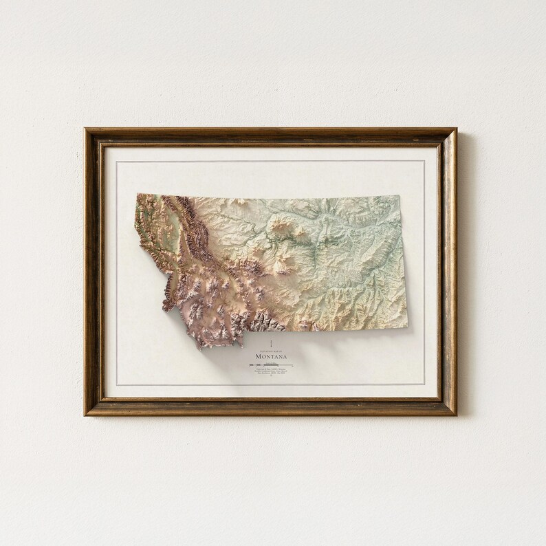 May include: Framed 3D relief map of Montana, showcasing the state's topography in shades of brown, green, and purple. The map is set against a white background and enclosed in a dark brown wooden frame. The word "Montana" is visible at the bottom.