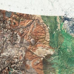 May include: A detailed topographical map with a blend of earthy tones, including shades of brown, beige, and green. The map highlights mountainous regions and river systems, with a portion of a white and gray map in the upper right corner.