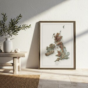 May include: Framed 3D relief map of the British Isles, showcasing the topography with shades of brown and green. The map is in a gold-colored frame, leaning against a white wall. A small wooden bench and a vase with greenery are in the foreground.