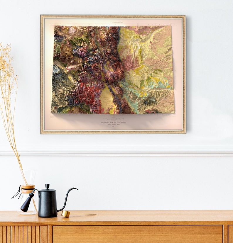 May include: A framed geological map of Colorado, featuring a detailed relief with various colors representing different geological formations. The map is in a gold-colored frame and hangs on a white wall. The text "GEOLOGICAL MAP OF COLORADO" is visible.