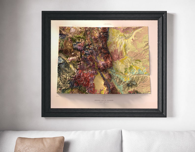May include: Framed geological map of Colorado, featuring a detailed relief with a variety of colors including red, green, yellow, and brown. The map is set in a black frame and mounted on a light pink background. The title "GEOLOGIC MAP OF COLORADO" is visible.