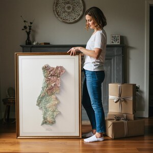 May include: A framed 3D relief map of Portugal, featuring a detailed topographical representation in shades of green, brown, and pink. The map is set against a white background within a gold-colored frame. The word "PORTUGAL" is printed at the bottom.
