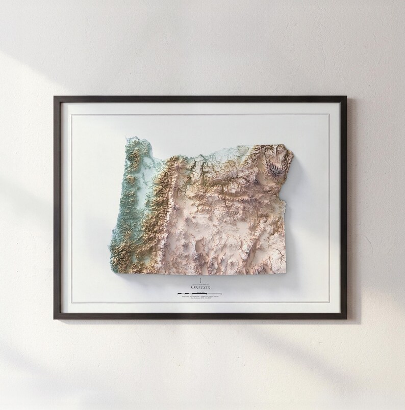 Puede incluir: Un mapa topogr&aacute;fico 3D enmarcado de Oreg&oacute;n, que muestra el terreno del estado con tonos de verde, marr&oacute;n y beige. El mapa est&aacute; sobre un fondo blanco dentro de un marco negro. La palabra "OREGON" est&aacute; impresa en la parte inferior.