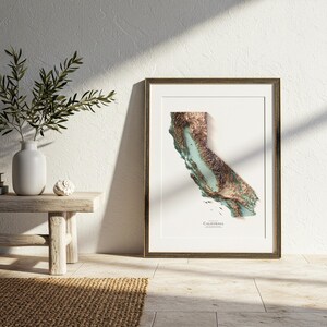 Op de afbeelding: Ingelijste 3D-reli&euml;fkaart van Californi&euml;, met de topografie van de staat in bruin, groen en blauw. De kaart staat tegen een witte achtergrond, met het woord "California" onderaan.