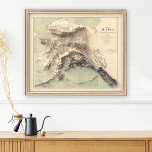 Framed custom Alaska map art displayed in a home office with a leather desk chair, vintage compass, and trail maps. The print focuses on the Inside Passage and Wrangell-St. Elias glaciers, inspiring wanderlust for outdoor enthusiasts or as a meaningful gift for someone planning their next Alaskan kayaking or fishing expedition.